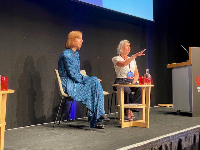 Two women are sitting on a stage addressing conference attendees with a small round table between them