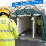 Image shows the back of a member of a FRS member dressed in high vis uniform and helmet standing on a road, looking at team members in a pop up tent.